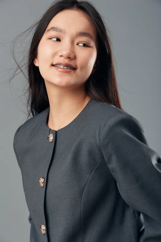 Young woman with braces smiling and looking sideways wearing a dark gray buttoned jacket against gray background, casual portrait of cheerful Asian female with long hair.