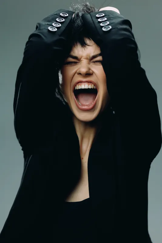 screaming woman in black jacket expressing strong emotions with closed eyes and raised hands, dark background