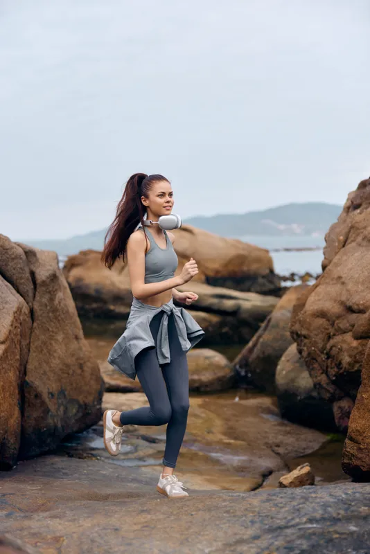 Young athletic woman jogging along rocky beach wearing sportswear, headphones, showcasing fitness and determination against a serene coastal backdrop