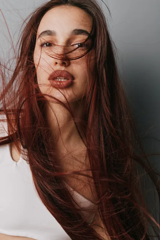 Woman with long hair blowing in wind wearing white top looking at camera with neutral expression on gray background. Portrait of young female with natural makeup and loose hair strands.