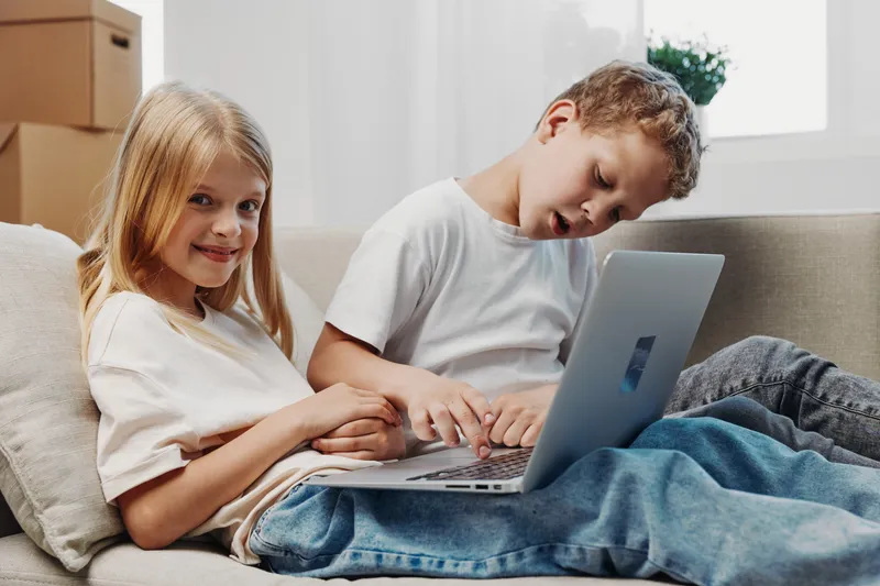 Two children sitting on sofa using laptop together in bright room. Girl smiling at camera while boy focuses on screen. Concept of learning, technology, childhood and home environment.