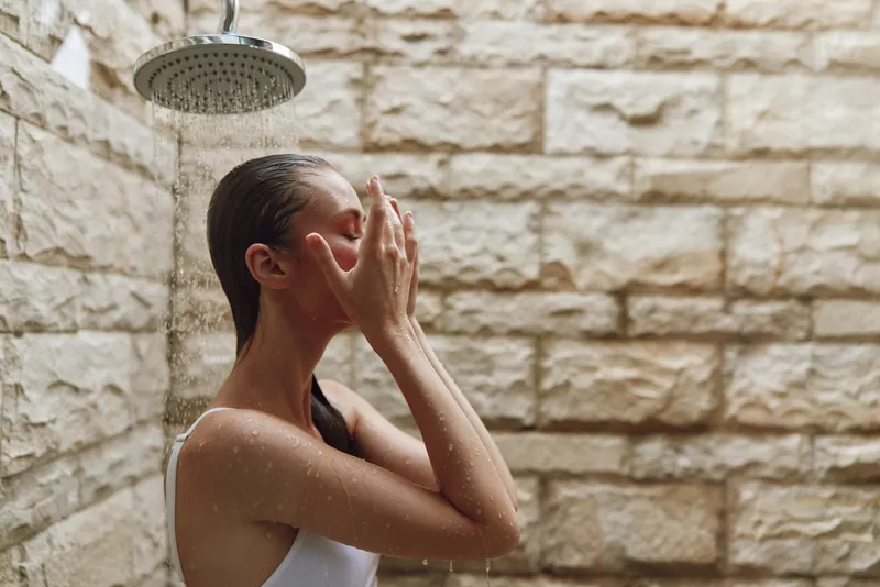 Woman washing face under outdoor shower with water drops, fresh skincare routine, natural stone wall background, serene wellness moment, healthy lifestyle.