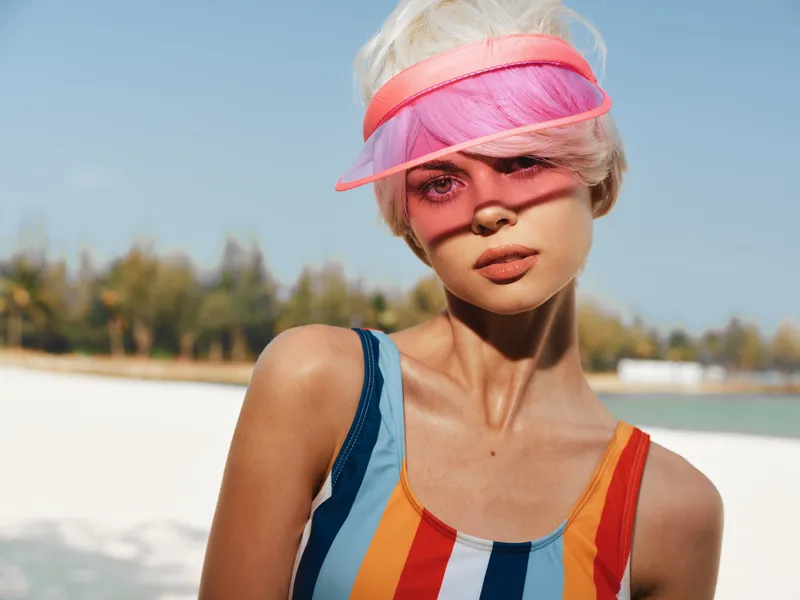 Close portrait of a sunlit woman at the beach wearing a vibrant striped swimsuit and pink visor, warm glow, clear sky, wind in hair, summery mood.