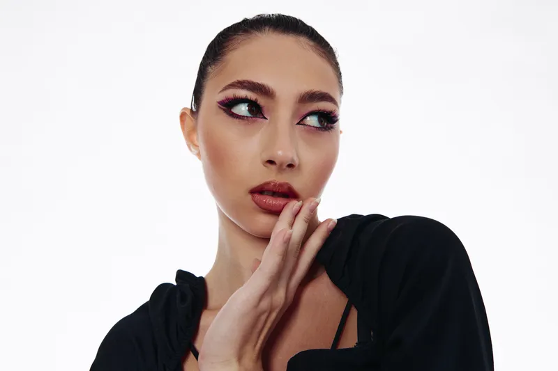 Fashionable woman with artistic makeup shows creative eyeshadow look, wearing black clothing. Studio portrait on white background, concept of beauty, fashion, and creative style.