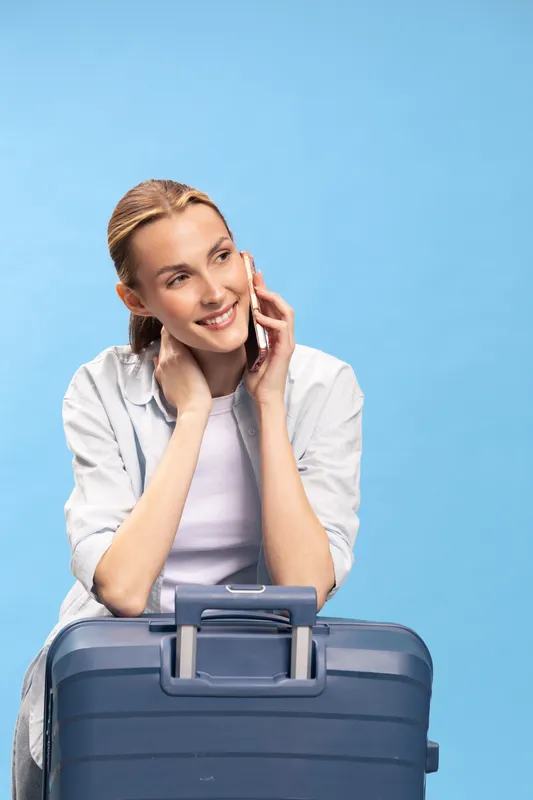 Smiling woman with suitcase talking on phone against blue background. Concept of travel, communication, vacation, and happiness in casual outfit with natural lighting.