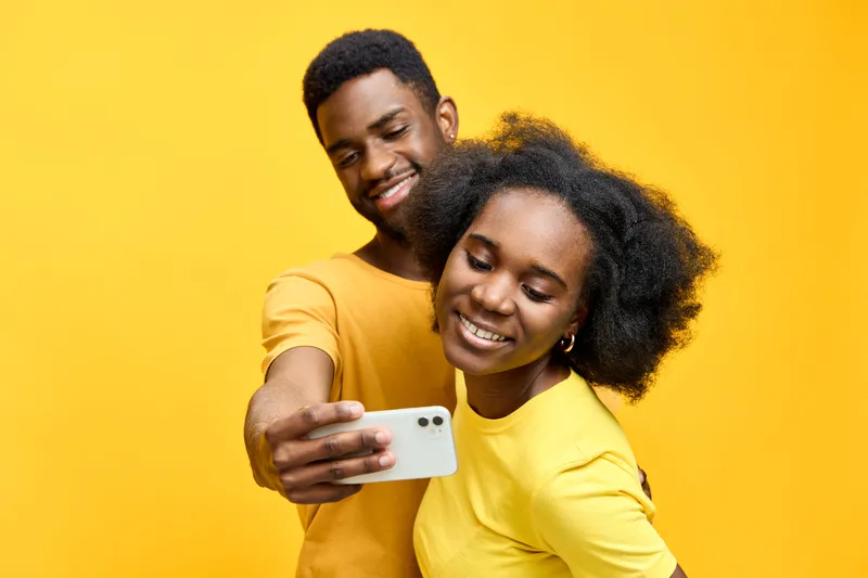 Young cheerful African couple taking a selfie with a smartphone in vibrant yellow shirts against a bright yellow background, capturing joyful emotions