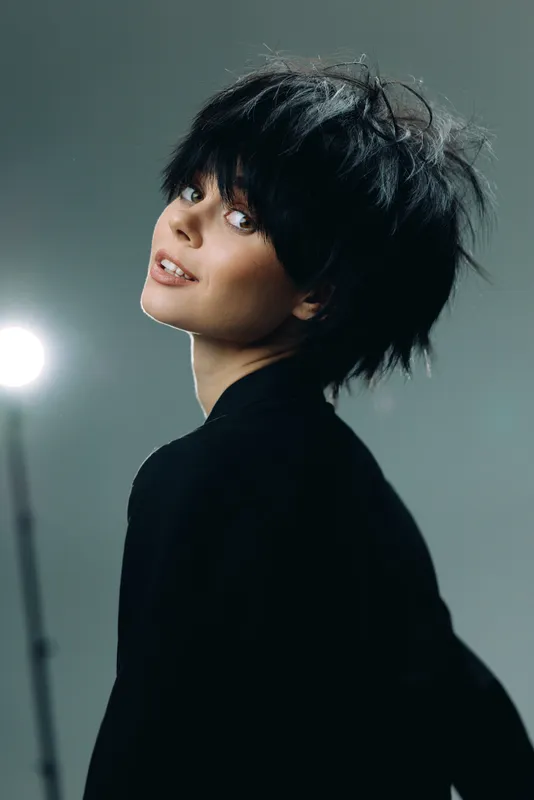 Smiling woman with short black hair and black clothing posing indoors with soft lighting and neutral background.