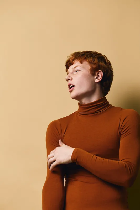 Young man with red hair wearing brown turtleneck sweater standing against beige background looking sideways with thoughtful expression and crossed arms.