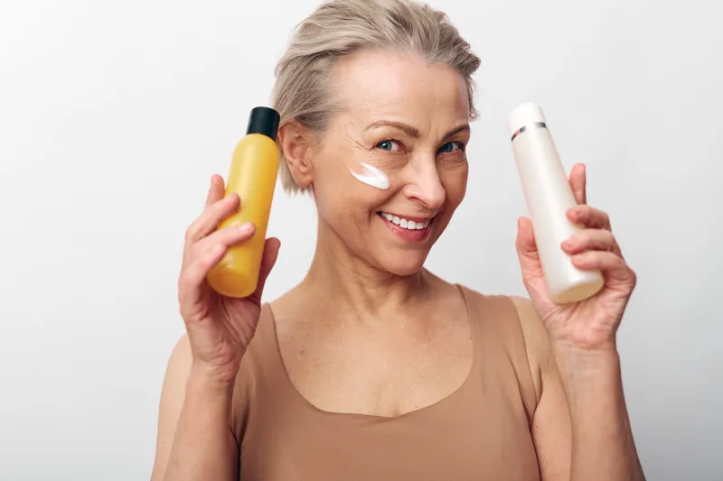 Smiling mature woman with short hair holds two skincare bottles, applying cream on her cheek. Concept for beauty routine, anti aging, hydration, and face care with natural cosmetics.