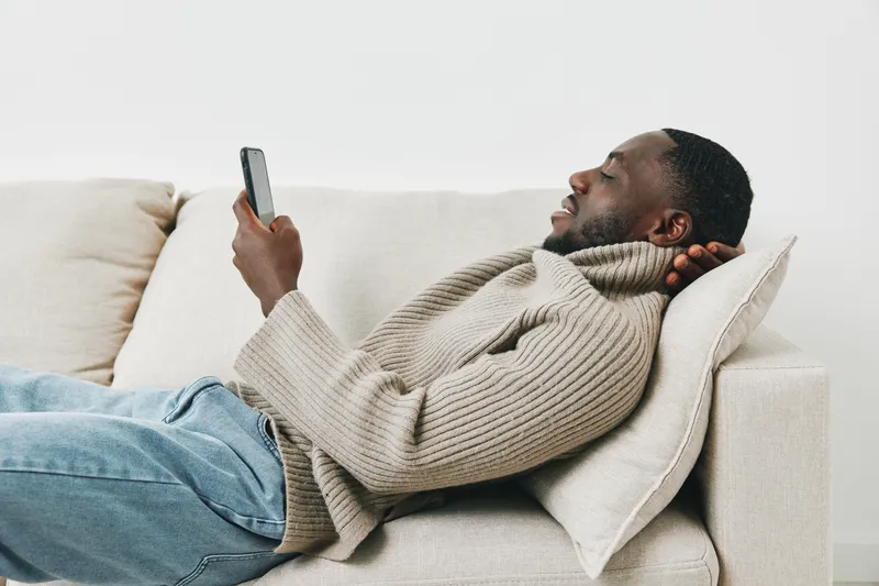 Young African man relaxing on a sofa while using a smartphone, casual outfit, cozy atmosphere, modern interior, light colors, and joyful expressions