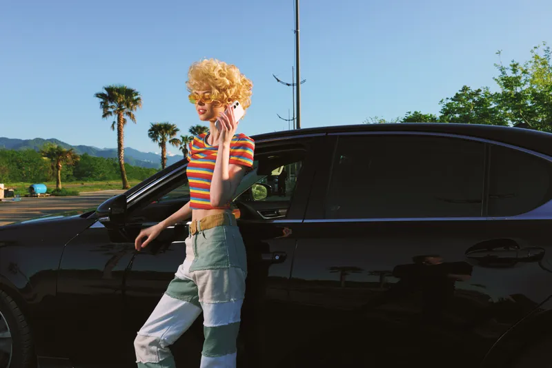 Curly blonde woman talking on a cellphone beside a dark car in bright outdoor light. Retro color palette, casual street style, and a confident mood evoke leisure and glamour.