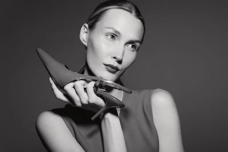 White woman with pale skin holds a high heel shoe near face in black and white studio portrait. Concept of fashion, elegance, and style.