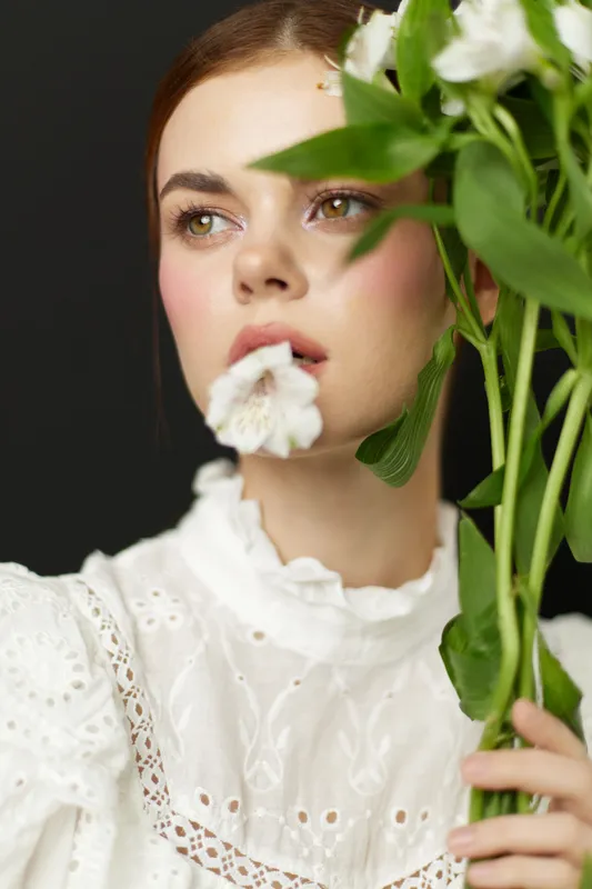 A contemplative young woman in a white vintage dress holds fresh flowers, showcasing natural beauty and emotional depth against a dark backdrop