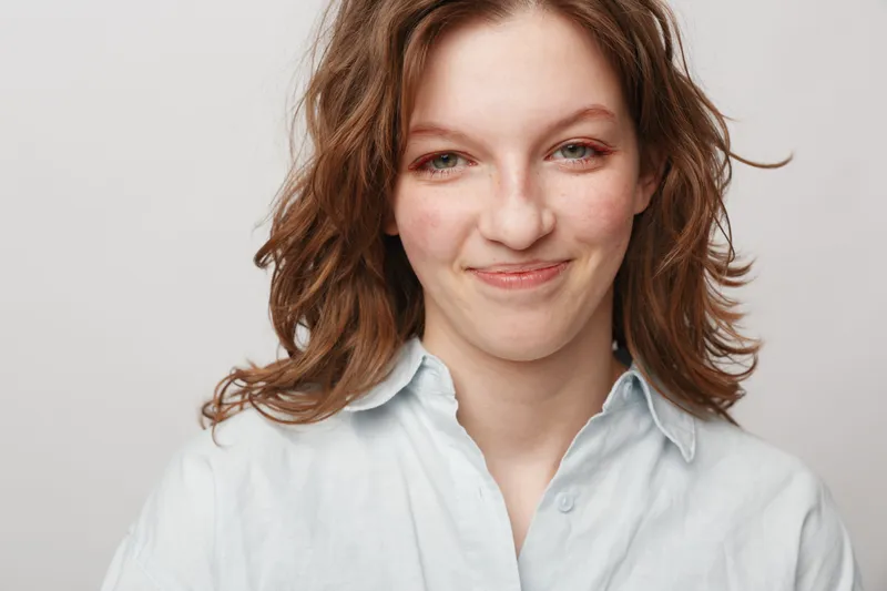 Young woman with wavy brown hair smiles gently wearing a white shirt against a plain light background. Concept of natural beauty, casual style, and positive emotions.