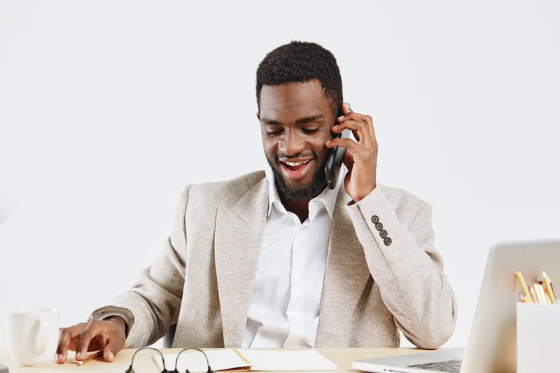Professional businessman in beige blazer working at desk with headphones on, smiling and talking on phone, using laptop and notepad in bright office space, modern corporate environment, joyful work mo