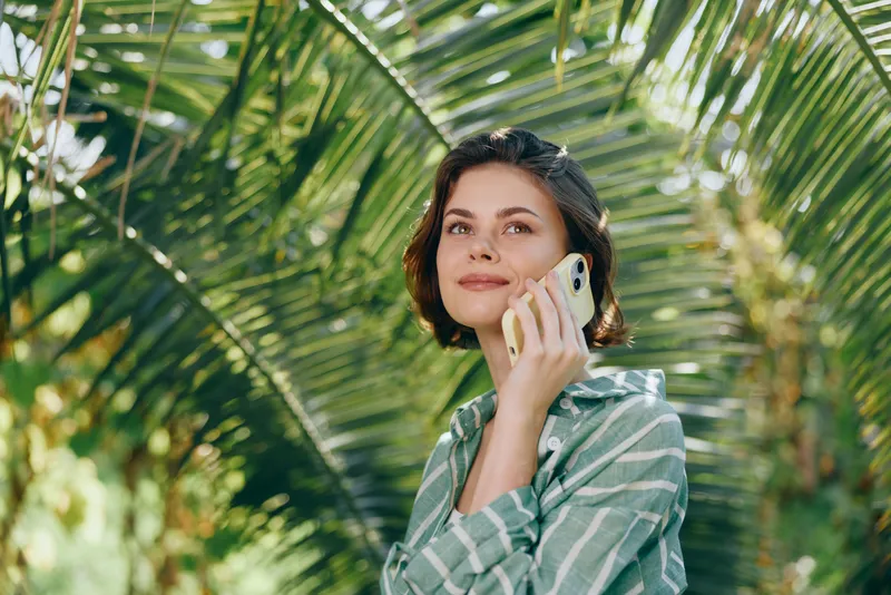Woman outdoors talking on a smartphone amid lush tropical foliage, wearing a striped shirt, soft sunlight, relaxed mood, natural colorful backdrop.