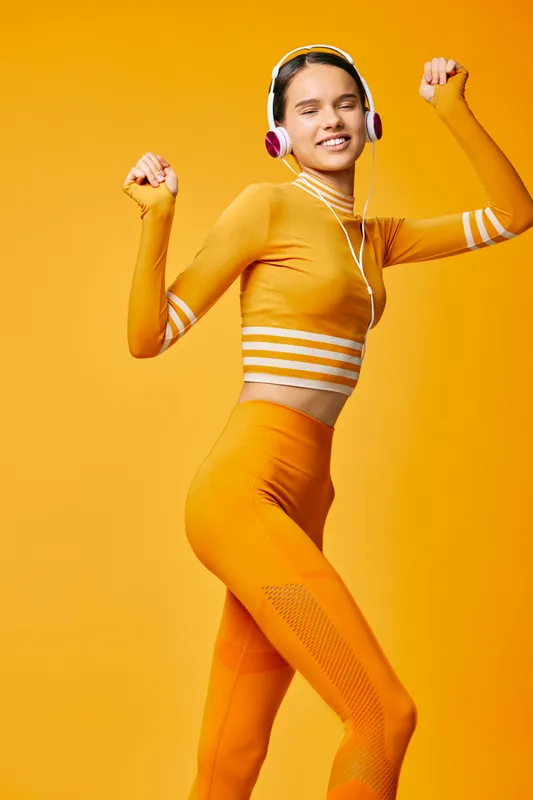 Fitness woman in a vibrant orange workout set with headphones, dancing energetically against a bright yellow background. Dynamic movement, cardio exercise, and upbeat studio vibe.