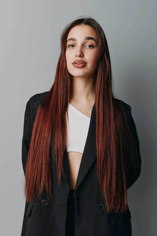 Young woman with long straight red hair wearing black blazer and white top standing against gray background looking confident and stylish with natural makeup and relaxed posture