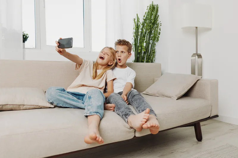 Two happy children sitting on couch taking selfie with smartphone in bright modern living room. Concept of childhood fun, friendship, and casual home moments.
