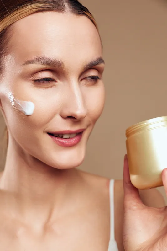 A smiling woman with clear skin applies white cream on her cheek while holding a gold cosmetic jar. The neutral background highlights skincare and beauty concepts for daily routine.