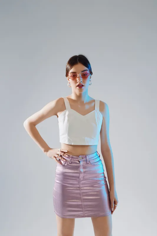 Young woman in a white crop top and shiny skirt poses confidently against a plain background. Fashion and beauty concept with modern style and attitude.