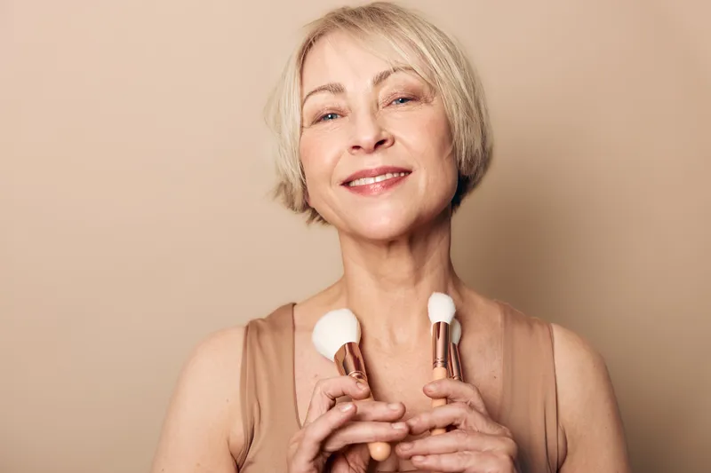 Smiling middle aged woman with short blonde hair holds two makeup brushes close to chest on beige background. Concept for beauty, skincare, cosmetics, self care, and mature woman confidence.