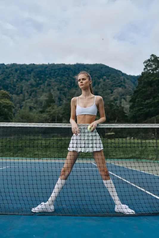 Athletic female model posing confidently on a tennis court, dressed in a white crop top and skirt, showcasing a healthy lifestyle amidst a natural backdrop