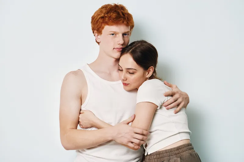 Young couple embracing each other with eyes closed and calm expression. Man with red hair and woman hugging, both dressed in casual white tops against plain background.