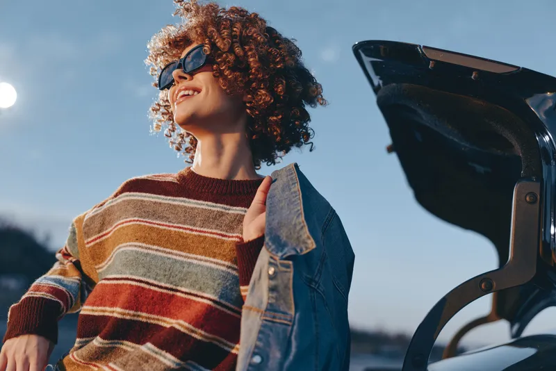 Woman lifestyle outdoors, curly hair, sunglasses, striped sweater and denim jacket, smiling in sunny sky near bicycle, relaxed street moment, looking upward in soft daylight now.