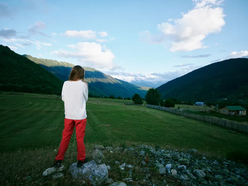 Woman standing on rocks overlooking a green valley with mountains in the distance, clear blue sky and scattered clouds, capturing a serene outdoor landscape scene with calm rural vibes.