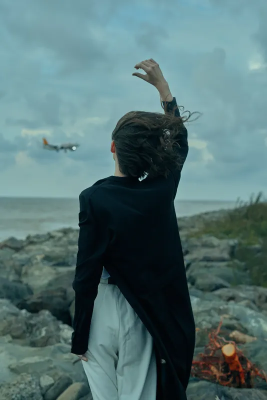 Ocean horizon behind a rocky shore, wind tousling a dark coat as a woman stands with back to viewer, hand raised toward the cloudy sky, conveying travel mood and dramatic coastline