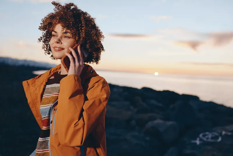Woman with curly hair holding phone outdoors at sunset, mountains behind her, warm evening light. Casual lifestyle portrait with relaxed smile, travel mood, natural atmosphere, soft blur.