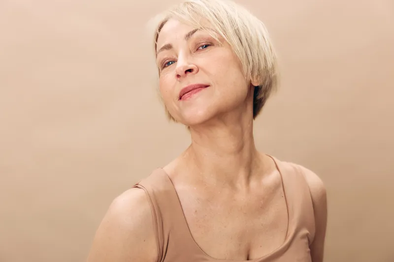 Confident middle aged woman with short blonde hair poses in beige sleeveless top against neutral background. Concept of natural beauty, aging gracefully, and self confidence.