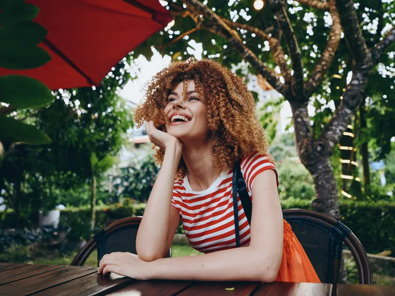 Portrait of a joyful woman with curly hair seated at a wooden outdoor table, wearing a striped red shirt, smiling under a bright umbrella on a sunny day.