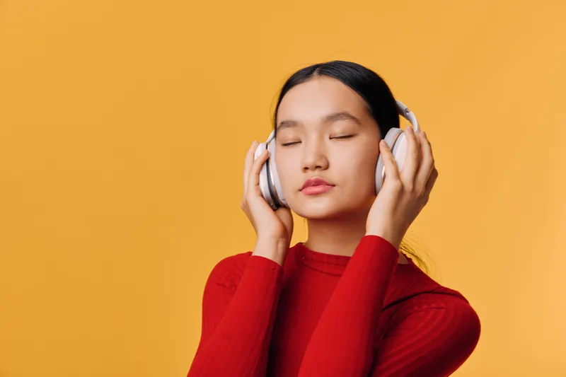 Young woman with closed eyes listening to music on wireless headphones, wearing red sweater on orange background, relaxing and enjoying sound in calm expression.