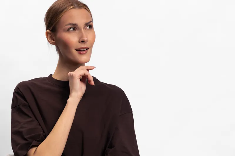 Thoughtful young woman in dark brown sweater looks sideways with hand near chin. Isolated on white background. Concept for contemplation, decision making, and curiosity in studio portrait.