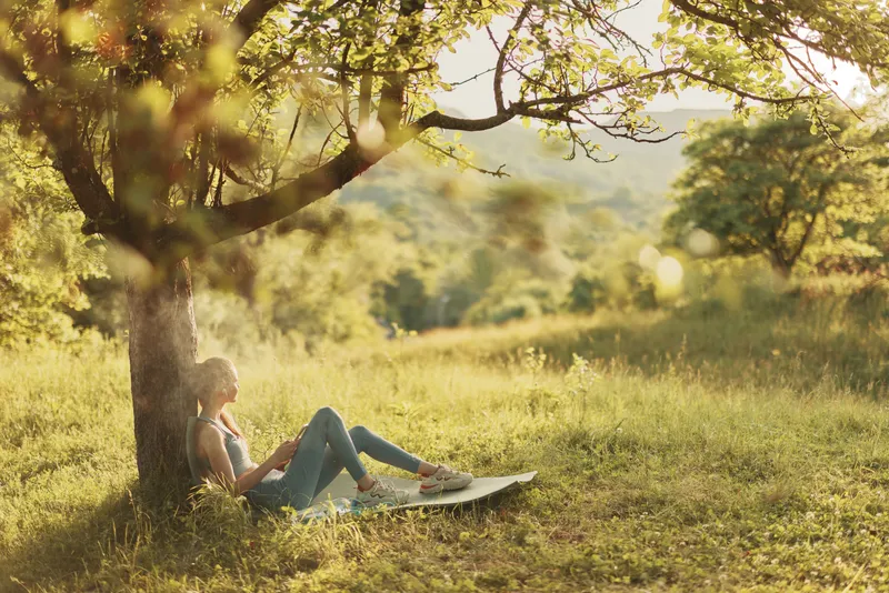Relaxed woman enjoying nature under a tree, surrounded by greenery, wearing casual blue attire, conveying peaceful emotions in a serene outdoor setting