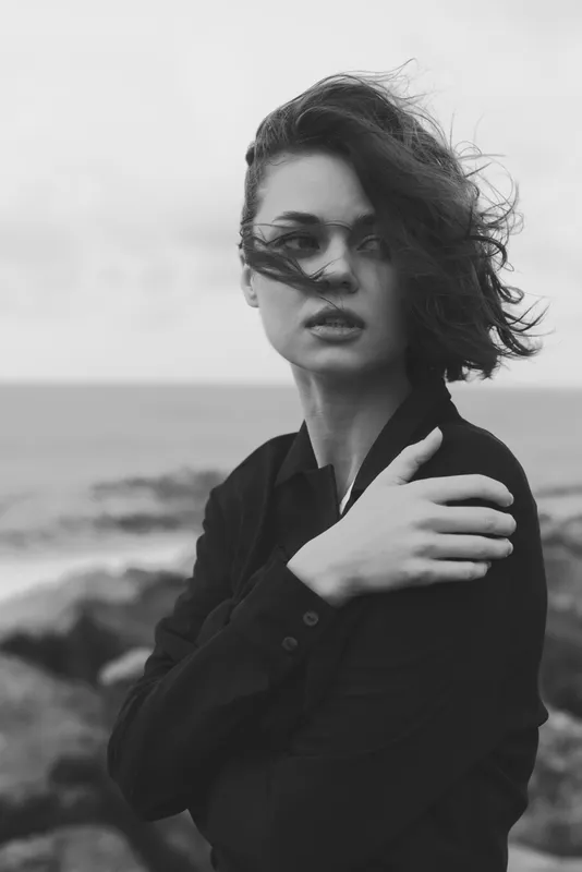 Black and white photo of a windblown woman by the sea, hair sweeping across her face, wearing a dark shirt, moody coastal atmosphere, monochrome fashion, dramatic silhouette and oceanic backdrop.