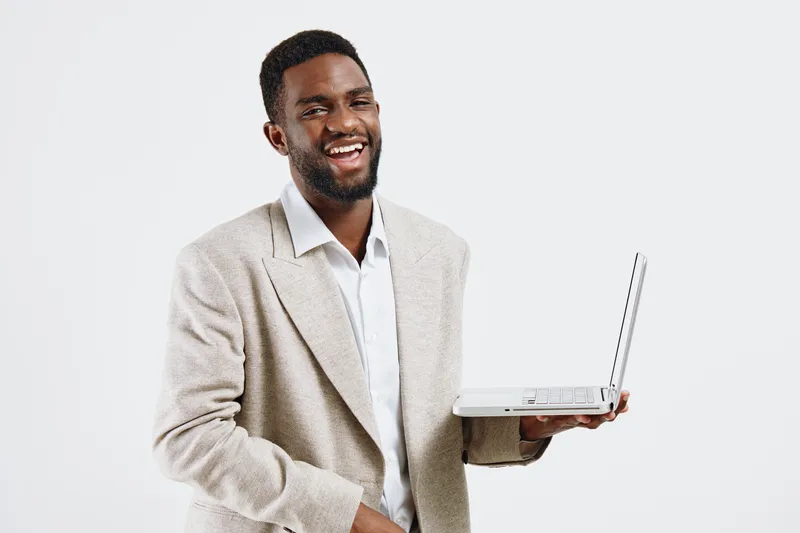 Happy smiling man of African descent wearing beige blazer and white shirt holding open laptop in front of plain white background. Business casual indoor portrait. People lifestyle concept