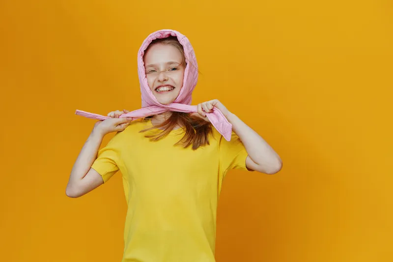 Happy girl wearing a pink scarf and yellow t shirt, smiling brightly against a vibrant yellow background, showcasing joy and playfulness