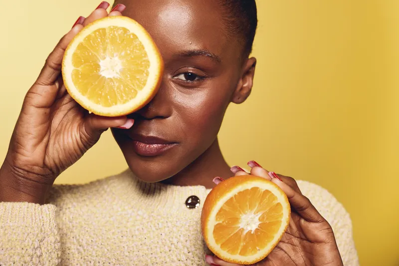 Close-up of a young black woman with dark skin holding two orange halves near her face against a yellow background. Concept of fresh fruit, healthy lifestyle, and natural beauty.