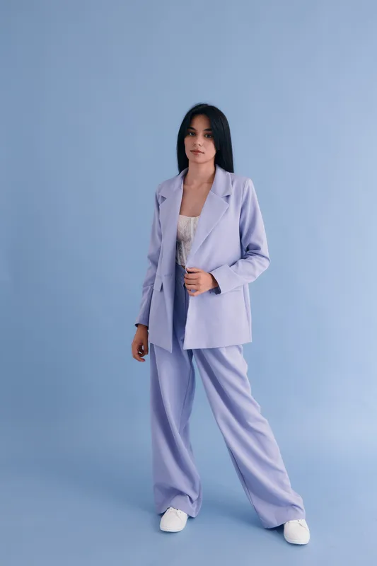 Young woman in lavender pantsuit stands against light blue background, wearing white sneakers with confident and calm expression, minimalistic fashion style.