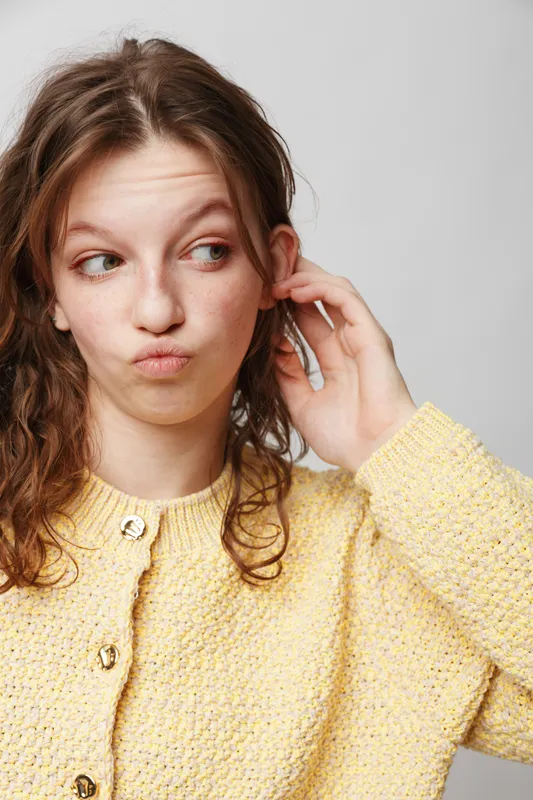 Young woman with curly hair wearing yellow knitted sweater looks to the side with thoughtful expression on light background. Concept of casual style, contemplation, and natural beauty.