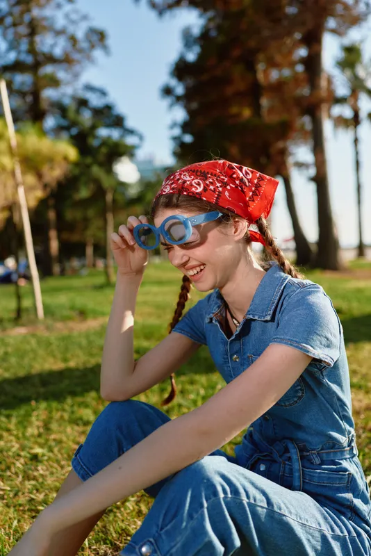 Lifestyle woman in modern boho-western denim street style with playful blue glasses and red bandana in sunny outdoor park, film color effect enhancing vibrant mood and fashion.