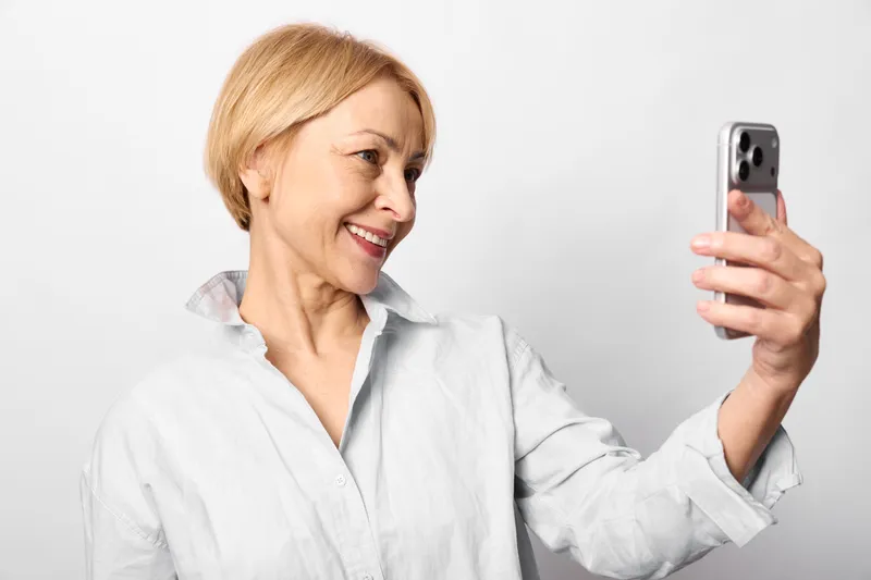 Woman selfie smartphone portrait, smiling older woman in studio with white background, holding smartphone for self portrait photo, closeup, casual