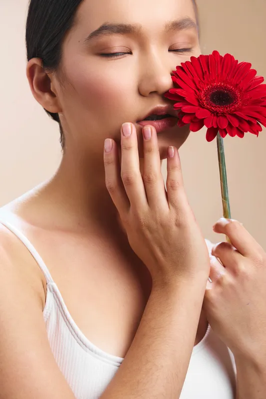 Asian woman softly holds a vibrant red flower near her lips, eyes closed, creating a tranquil beauty moment with warm light, smooth skin, and delicate texture.