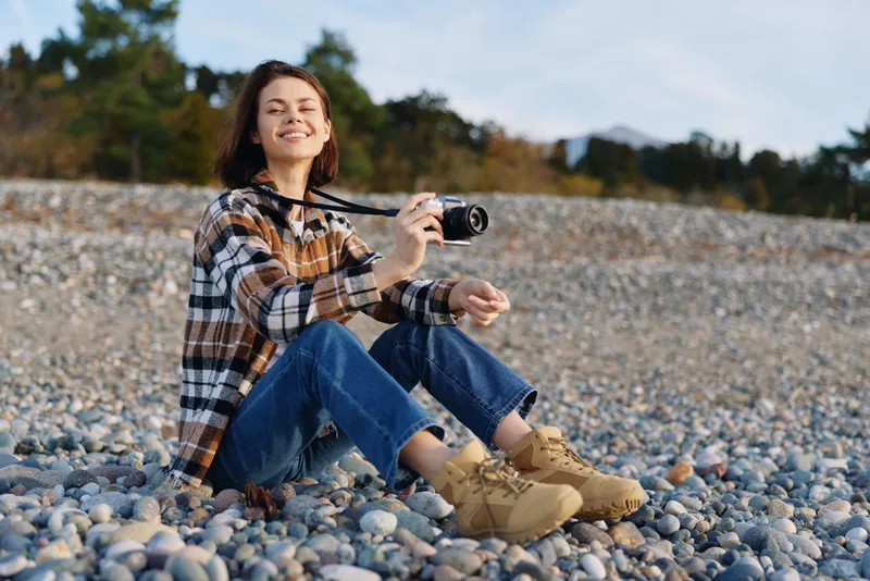 Woman photographer sits on a pebble beach, smiling while holding a camera. Casual plaid shirt, denim jeans, and boots showcase relaxed creativity in a sunlit outdoor setting with distant trees.