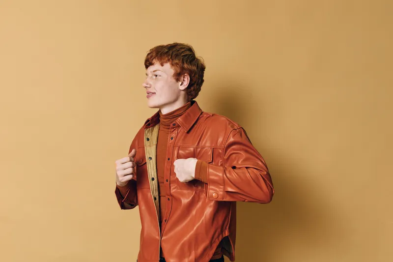 Young man with red hair wearing orange jacket and brown turtleneck standing against beige background in casual autumn outfit with confident posture and side profile view.