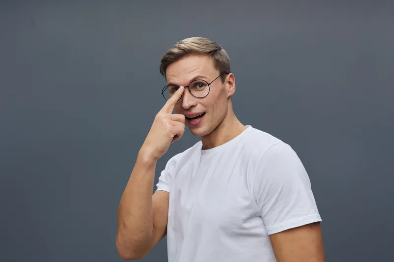 Young caucasian man in glasses white t-shirt touch face surprised emotional digital detox concept isolated gray background studio portrait