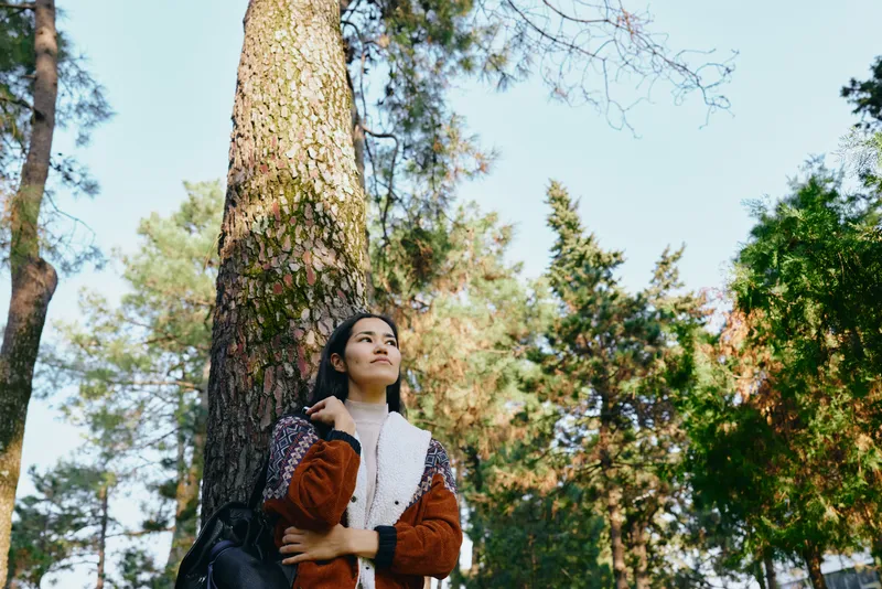 Forest, woman leaning against a tall tree, gazing upward in a serene moment of nature, outdoor reflection under dappled sunlight and calm woodland atmosphere.
